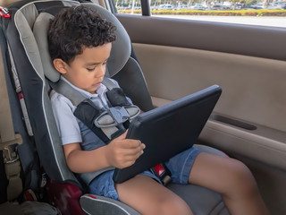 A little boy sets in the car seat playing with a tablet. Travel while entertaining the kids with smart apps on the tablet. School anywhere.