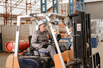 man driving a forklift through a warehouse in a factory. driver in uniform and protective helmet....
