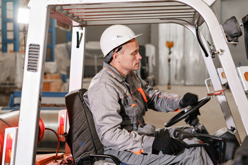 Naklejka premium man driving a forklift through a warehouse in a factory. driver in uniform and protective helmet. the concept of logistics and storage
