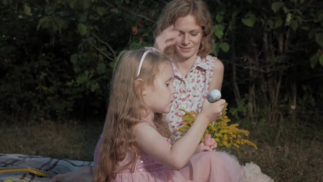A happy mother and daughter inflate soap bubbles. Family in a city park on a picnic on a warm evening at sunset.