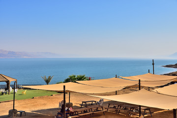 shore of the dead sea, canopies for protection from the sun, benches and tables for holidaymakers, sea horizon, haze over the sea, cloudless blue sky