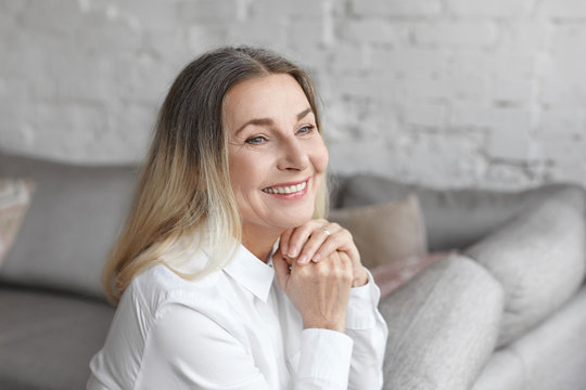 Positive human emotions, reaction, feelings and attitude. Gorgeous happy elderly European woman with loose hair and charming joyful smile, rejoicing at good positive news, sitting at home on couch