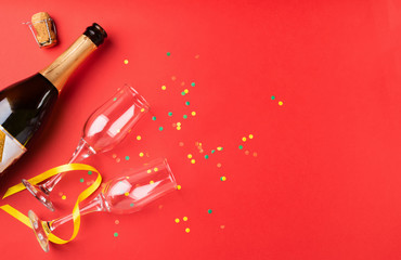A bottle of champagne and empty glass goblets are lined up on a red background decorated with confetti and serpentine. Festive concept. Flat layout.