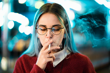 Millennial pretty girl with unusual dyed hairstyle smoking cigarette in amusement park at night....