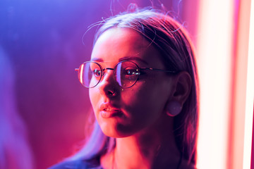 Millennial enigmatic pretty girl with unusual dyed hairstyle near glowing neon wall at night. Blue hair, golden sequins as freckles,nose piercing. Mysterious hipster teenager in glasses