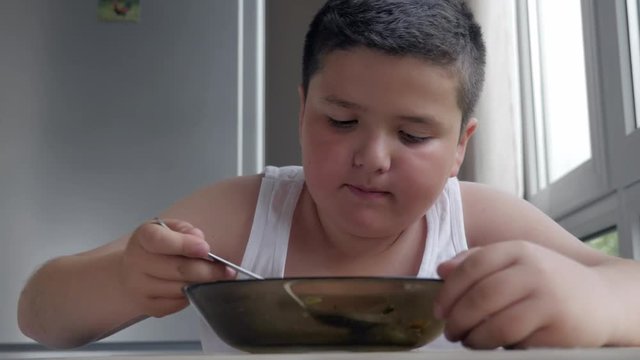 cute little fat boy dining in the kitchen, eating a tablespoon of soup, concept childhood obesity and gluttony