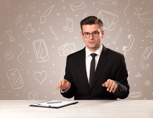 Young handsome businessman sitting at a desk with white mixed media icons behind him
