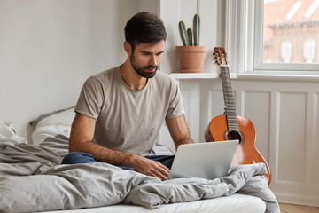 Attractive young bearded freelancer sits in front of opened laptop computer, watches tutorial video online, keeps legs folded, enjoys working process in comfortable bed, chats on modern notebook