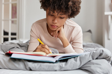 Peaceful atmosphere. Cropped shot of pleasant looking dark skinned woman writes notes in notebook...