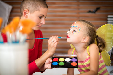 Fototapeta premium Young mother painting daughters face for Halloween party. Halloween or carnival family lifestyle background. Face painting and dressing up.