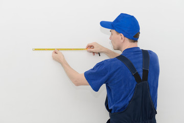 worker in blue overalls measures the distance on a white wall using a tape measure