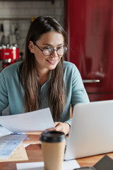 Delighted pleased young prosperous woman CEO creats successful plan for banking, works on creative business idea, holds paper documents, looks positively at laptop computer, sits at kitchen table.
