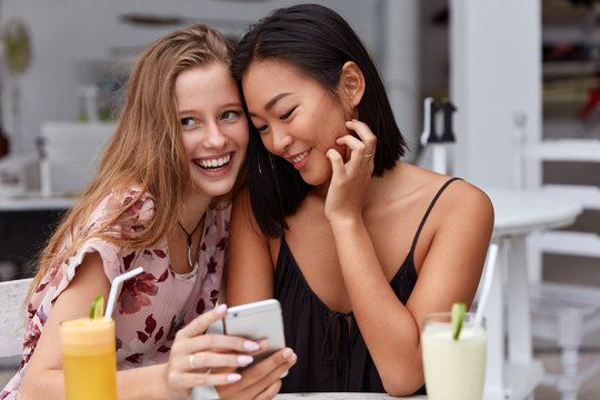 Mixed race young female friends read notification on smart phone, have positive facial expressions, drink fresh juice, pose indoor, enjoy online communication, sit together in cozy cafeteria. - Powered by Adobe