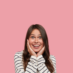 Amazed beautiful brunette woman keeps both hands on cheeks, has toothy smile, notices something...