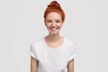 Adorable freckled young lady or teenager smiles joyfully at camera, has red hair combed in knot,...