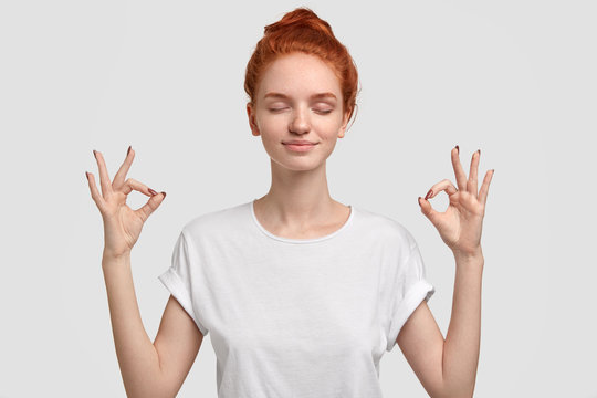 People, yoga and mediatation concept. Relaxed foxy girl with freckled soft skin enjoys peaceful atmosphere, keeps hands in mudra sign, relaxed after intense day, isolated over white background