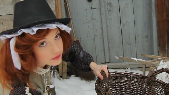 A beautiful red-haired girl in ethnic clothes is standing with a large basket on the street in winter. There is a domestic rat on the girl s hat. Fairy-tale character. Cinderella