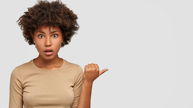 Studio shot of shocked dark skinned girl has puzzled indignant expression, points with thumb aside, talks about something terrible, dressed in casual sweater, isolated over white background.