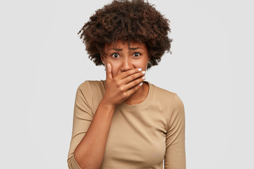 Studio shot of sad worried dark skinned girl has depressed expression, covers mouth with palm,...
