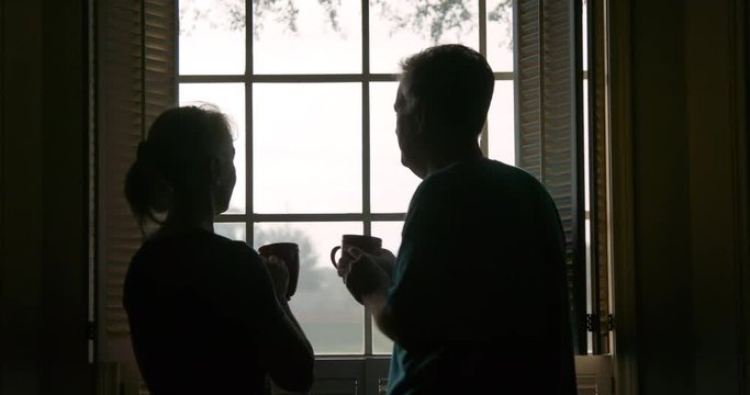 A man and a woman standing by a window looking out into suburbia drinking coffee on a leisurely weekend morning who are silhouetted by the daylight outdoors display a little affection.