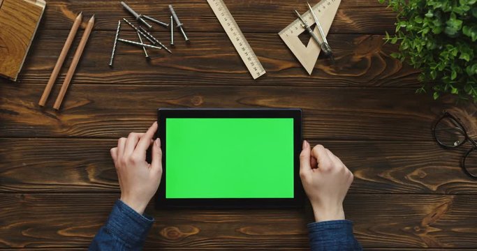 Top view on the black tablet device lying horizontally on the wooden table with office supplies and nails, female hands scrolling and tapping on the green screen. Chroma key.