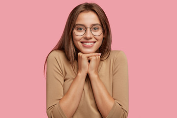 Photo of pleasant looking brunette Caucasian woman has toothy smile, holds chin with both hands,...
