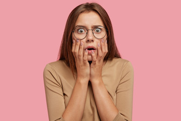 Half length portrait of beautiful female being very emotional and surprised, stares with bugged eyes, has stupor, dressed casually, isolated over pink background. Amazement and fear concept.