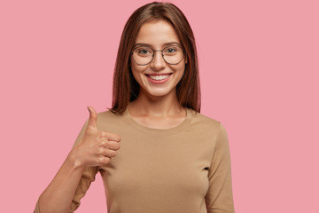 Portrait of happy teenager keeps thumb raised, being in good mood, shows her agreement, poses over pink background, says: Nice joke! Young woman shows like gesture, satisfied with something. - obrazy, fototapety, plakaty