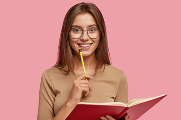 Photo of happy brunette woman with positive smile, carries textbook, holds pencil for writing,...