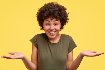 Uncertain clueless cheerful dark skinned lady in casual military t shirt, shruggs shoulders with...