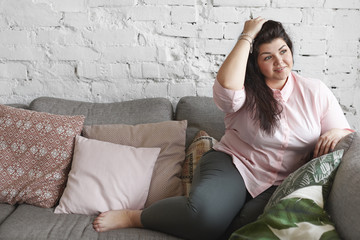 Rest, leisure and relaxation concept. Picture of well dressed young overweight female with chubby cheeks and curvy body sitting comfortably on gray couch against white brick wall background