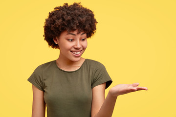 Studio shot of pleasant looking cheerful African woman raises palm near shoulder, holds copy space, smiles cheerfully, isolated over yellow background, dressed in casual t shirt. Advertisement