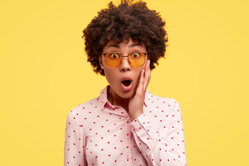 Photo of terrible young black woman keeps jaw dropped, holds hand on cheek, wears trendy...