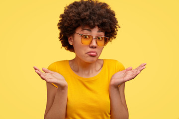 Uncertain dark skinned female shruggs shoulders with puzzlement, dressed in casual yellow t shirt, feels uncertain, makes decision, isolated over yellow background. Woman doesnt care, has no idea