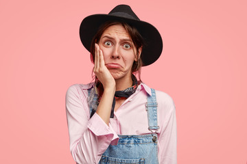 Horizontal shot of displeased female gardener purses lower lips, keeps hand on cheek, expresses...