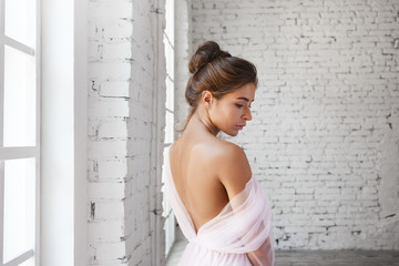 Attractive tender young woman with hair bun standing in spacious room wearing pink ball dress, demonstrating her bare shoulder. Slim beautiful bride with tanned perfect skin posing in wedding gown