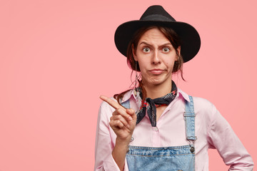 Dissatisfied female gardener frowns face in displeasure, points aside with index finger, shows direction aside, dressed in fashionable shirt, dungarees and hat, isolated over pink background.