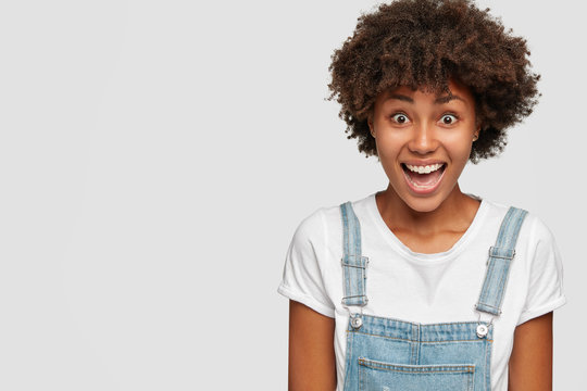 Indoor shot of cheerful dark skinned woman dressed in dungarees, laughs positively, can not hold excitement inside, being overwhelmed with happiness and positive emotions, isolated on white background