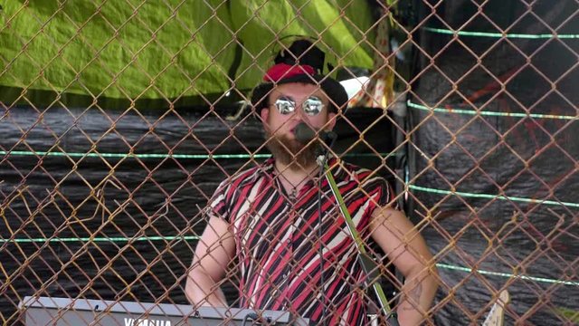 Grungy ringmaster commentates from behind a fence.