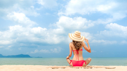 Woman in pink bikini with sun hat sitting at the beach in summer