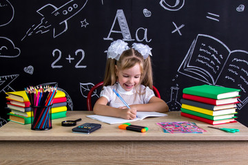 Excited and happy schoolgirl sitting at the desk with books, school supplies, holding the pen in...