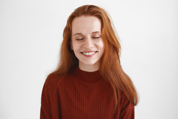 Fototapeta premium Cheerful young red haired woman with pleasant appearance closing eyes, smiling broadly, being in good mood, enjoying nice day, feeling relaxed and carefree. Ginger student girl expressing joy