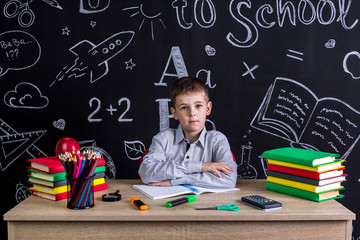 Serious excellent schoolboy sitting on the desk with books, school supplies, with both arms leaned one to another