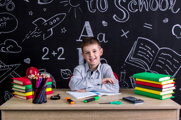 Happily smiling excellent schoolboy sitting on the desk with books, school supplies, with both arms leaned one to another
