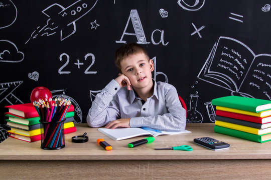 Puzzled excellent schoolboy sitting on the desk with books, school supplies, with one arm leaned under the cheeck