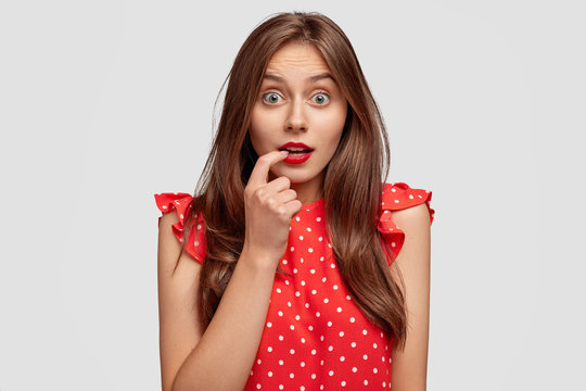 Omg, why it happens with me? Good looking brunette woman has dark hair, stares with bugged eyes, bites finger, dressed in polka dot blouse, cant believe in shocking news, isolated on white wall