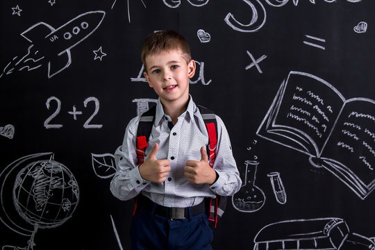 Smiling schoolboy standing before the chalkboard as a background with a backpack on his back with thumbs raised up. Lucky to go school again. Landscape picture