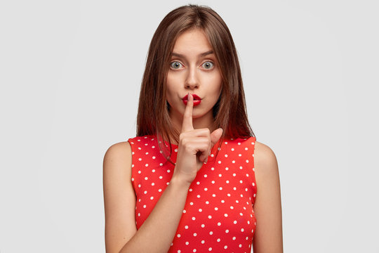 Pleasant looking long haired young woman wears red lipstick, makes silence gesture, asks not tell anyone secret, forbids talk loudly, dressed in polka dot dress, isolated over white background.