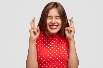 Pleased attractive European woman makes wish to win, raises hands with crossed fingers, waits for lottery results, closes eyes, has red lips, dressed in fashionable dress, isolated over white wall