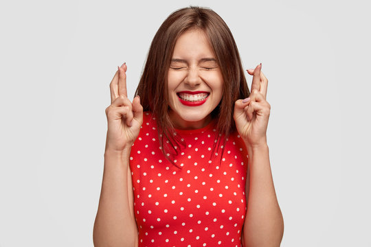 Pleased attractive European woman makes wish to win, raises hands with crossed fingers, waits for lottery results, closes eyes, has red lips, dressed in fashionable dress, isolated over white wall